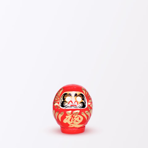 A 9-centimeter tall red Fuku-iri lucky daruma doll, made by Imai Daruma Naya, featuring a rotund body and decorative patterns for eyebrows and beard and gold body lines and kanji lettering.