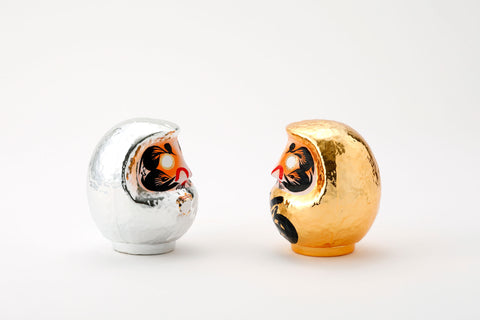 A pair of facing shiny Platinum Daruma lucky dolls — one silver and one gold — made by Imai Daruma Naya. Both feature rotund bodies and decorative patterns for eyebrows and beards. The silver has kanji lettering in white, the gold has lettering in black. 