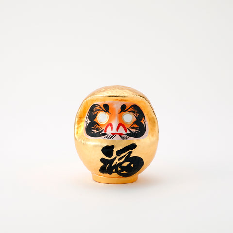 A shiny, gold Platinum Daruma lucky doll, made by Imai Daruma Naya. The rotund doll features decorative patterns for its eyebrows and beard, and black kanji lettering.