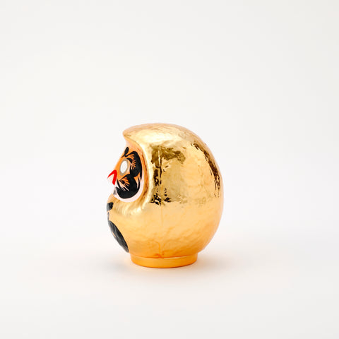A side view of a shiny, gold Platinum Daruma lucky doll, made by Imai Daruma Naya. The rotund doll features decorative patterns for its eyebrows and beard, and black kanji lettering.