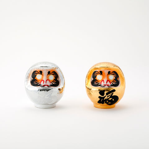 A pair of shiny Platinum Daruma lucky dolls — one silver and one gold — made by Imai Daruma Naya. Both feature rotund bodies and decorative patterns for eyebrows and beards. The silver has kanji lettering in white, the gold has lettering in black. 