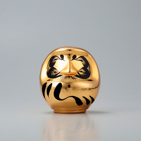 A shiny, gold Platinum Designer’s Daruma lucky doll, made by Imai Daruma Naya. The rotund doll features decorative patterns for its eyebrows and beard, and black curvy lines across its body.