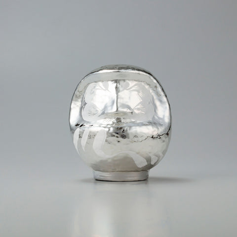 A shiny, silver Platinum Designer’s Daruma lucky doll, made by Imai Daruma Naya. The rotund doll features decorative patterns for its eyebrows and beard, and white curvy lines across its body.
